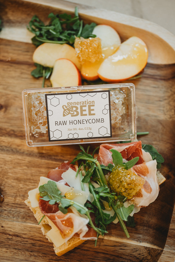 A clear plastic container of Generation Bee  raw honeycomb on a wooden table, surrounded by prosciutto, cheese, and apple slices
