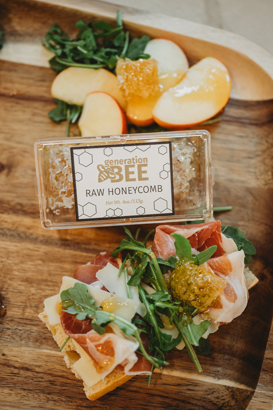 A clear plastic container of Generation Bee  raw honeycomb on a wooden table, surrounded by prosciutto, cheese, and apple slices