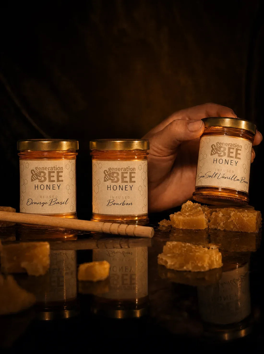 Three jars of Generation Bee infused honey on dark glass, with honeycomb and a honey dipper beside them